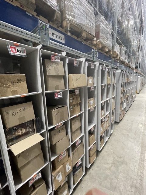 Rows of shelves in a warehouse filled with cardboard boxes, some labeled with numbers, under a high ceiling with pallets stacked above.