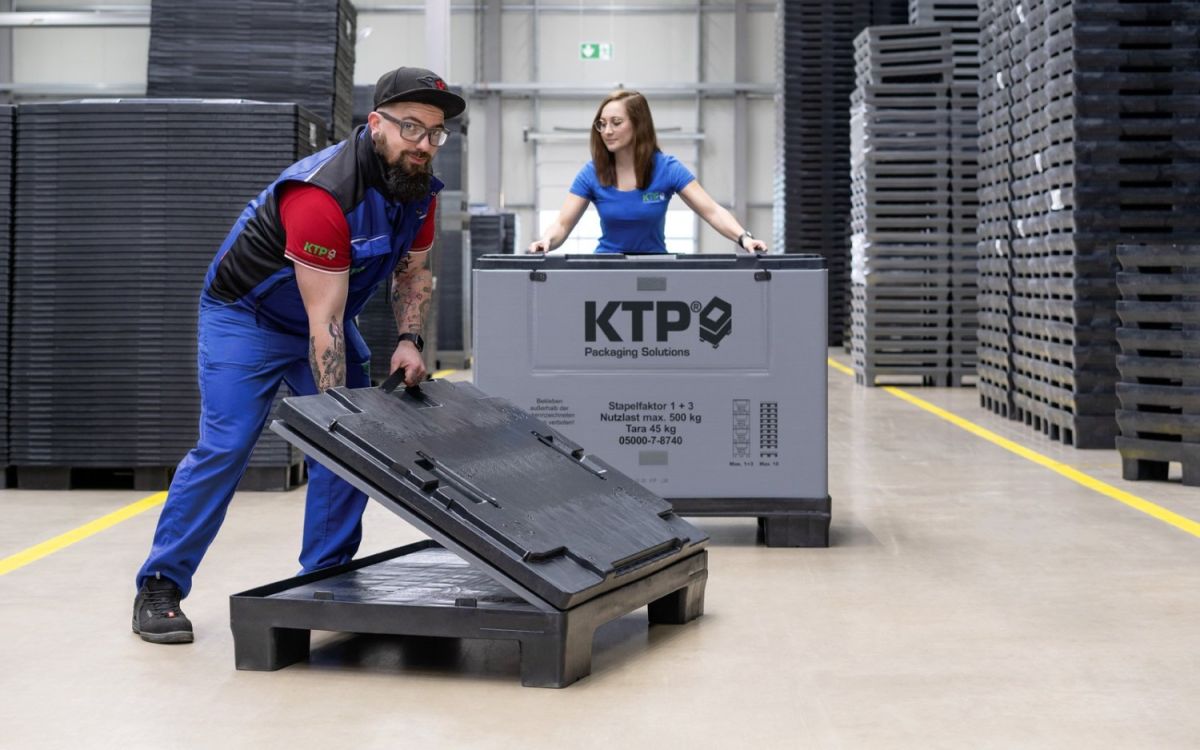 A man with a beard in a blue and black work uniform lifts a plastic lid while a woman in a blue shirt stands behind him next to a large grey packaging crate in a warehouse setting filled with stacked pallets.