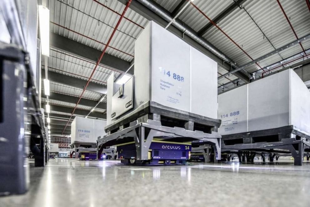 Automated guided vehicles transporting pallets in a warehouse with high ceilings and industrial lighting.