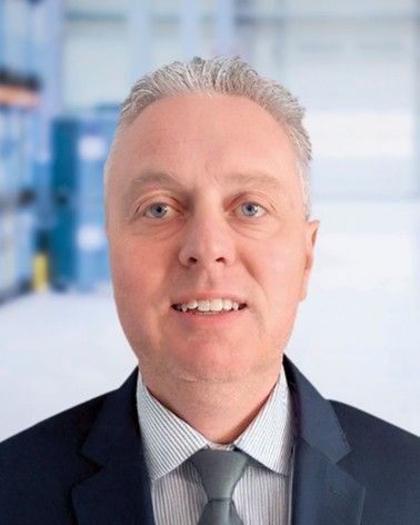 A man wearing a suit and tie, smiling, with a blurred industrial background.