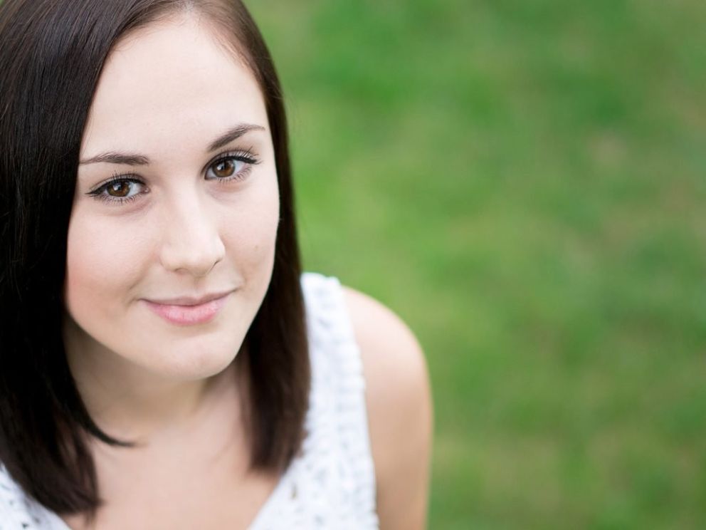 A young woman with long brown hair, wearing a white top, smiles softly against a blurred green background.
