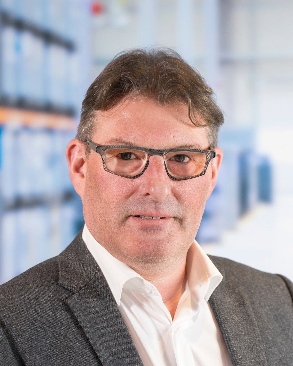 A professional man with short hair and glasses, wearing a grey blazer over a white shirt, poses in front of a blurred background that suggests a warehouse or storage setting.