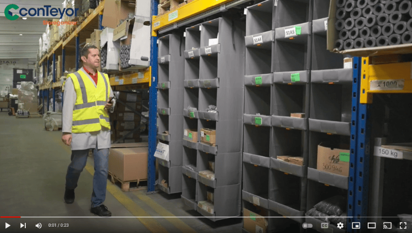 A warehouse scene featuring a man in a safety vest walking past organized shelves with labeled bins and boxes.
