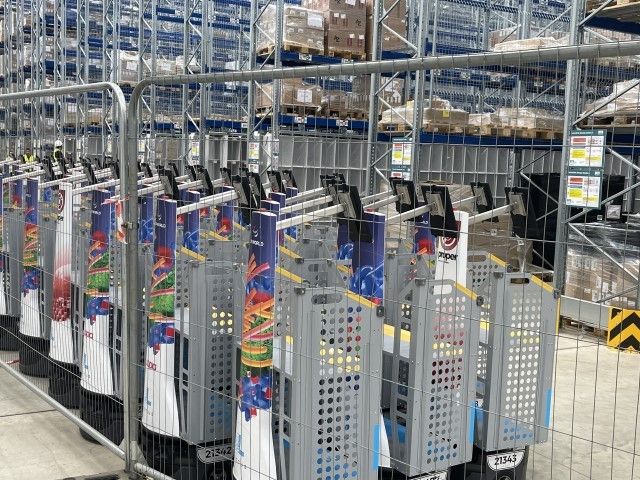 A row of self-service kiosks in a warehouse setting, surrounded by metal fencing and stocked shelves in the background.