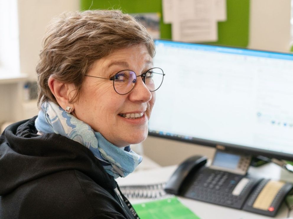 A woman with short, wavy hair and glasses smiles at the camera while sitting at a desk with a computer and a phone in the background. She is wearing a black hoodie and a light blue scarf.