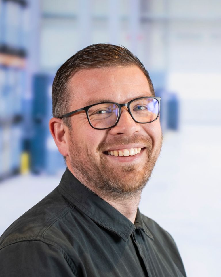 A smiling man with glasses and a beard, wearing a black shirt, set against a blurred background.