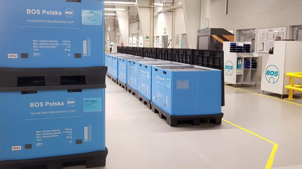 A row of blue shipping containers labeled "BOS Polska" in a manufacturing facility, with yellow safety striping on the floor and shelving in the background.