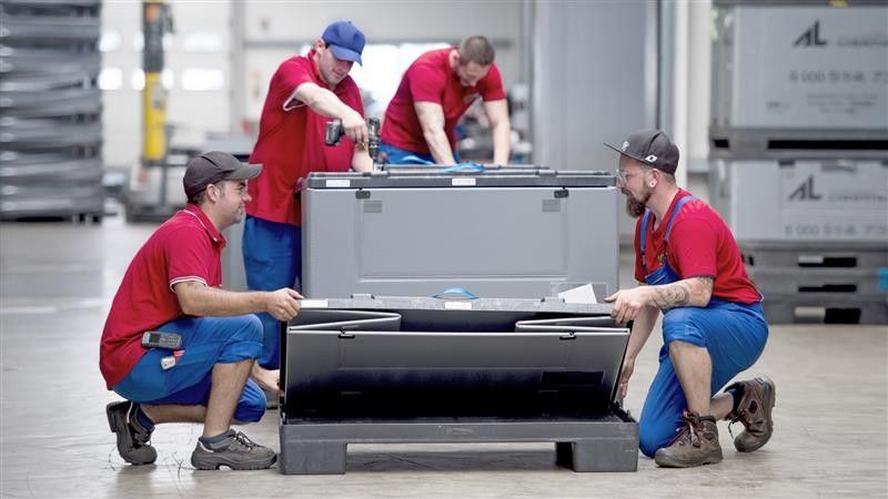 Four workers in red shirts and blue overalls are assembling a large container in a warehouse setting.