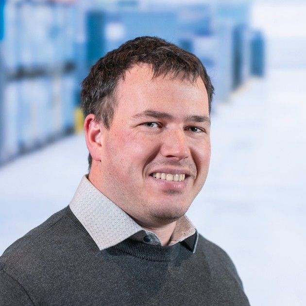 A smiling man in a grey sweater with a collared shirt, set against a blurred background of storage units.