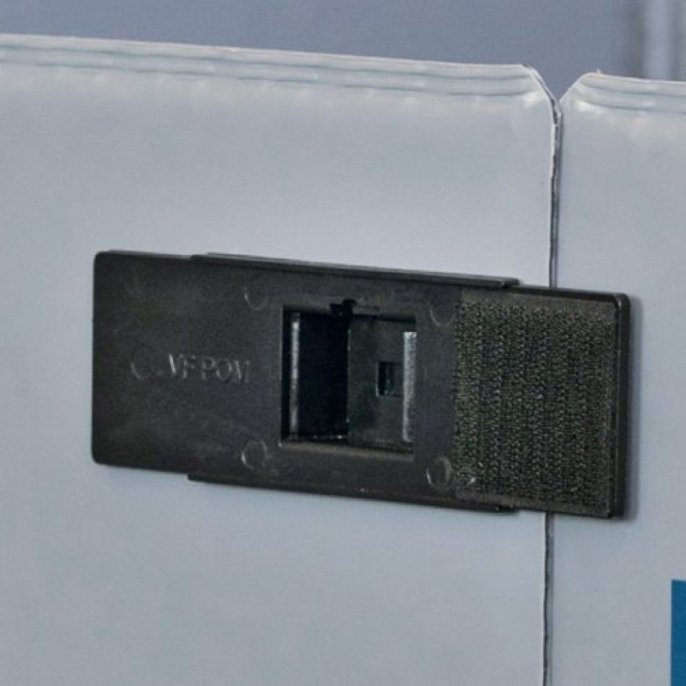A close-up image of a black plastic rectangular fastener attached to the edge of a white object, featuring a hook-and-loop strip on one side and a slot for securing items.