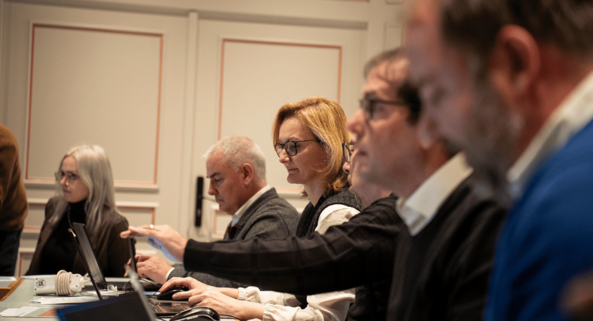 A group of people seated at a table, engaged in discussion or work, with laptops and documents in front of them.