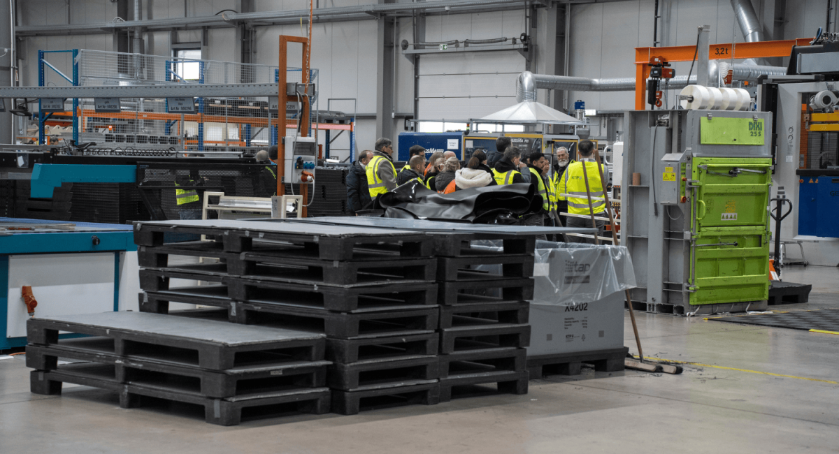 A group of people in safety vests gathered around a manufacturing area with pallets and machinery in a factory setting.