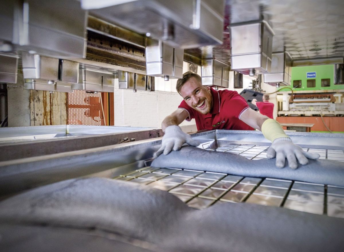 A smiling worker in a red shirt using a machine in a manufacturing setting, handling materials with gloves and surrounded by industrial equipment.
