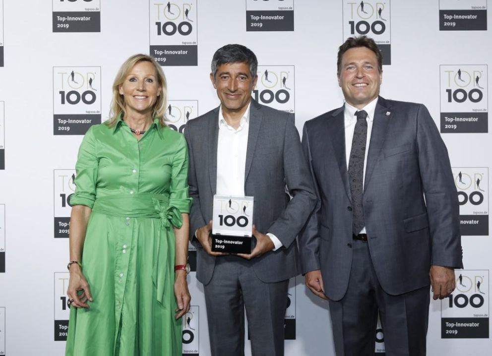 Three people posing for a photo at the Top Innovator 2019 event, with one person holding a trophy that reads "Top Innovator 100." The background features logos related to the event.