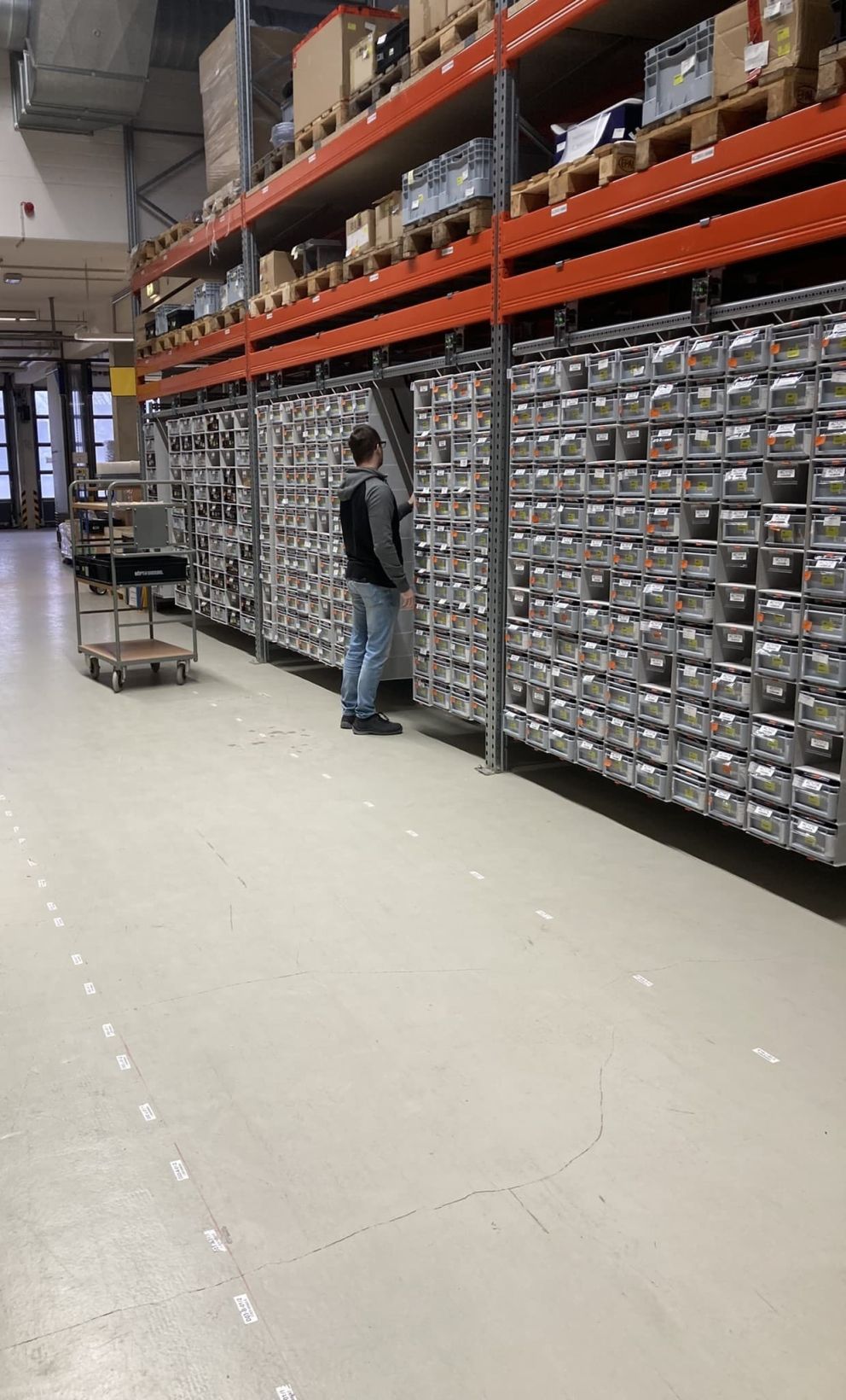 A person standing in a warehouse aisle, looking at shelves filled with labeled storage bins, with a cart in the foreground.