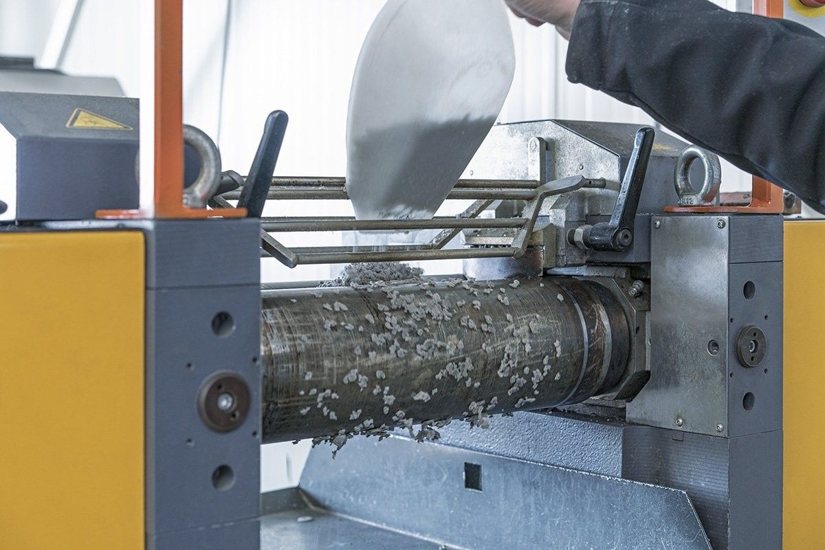 A worker using a spatula to apply material into a processing machine, with a focus on the machine's cylindrical roller and metal components.