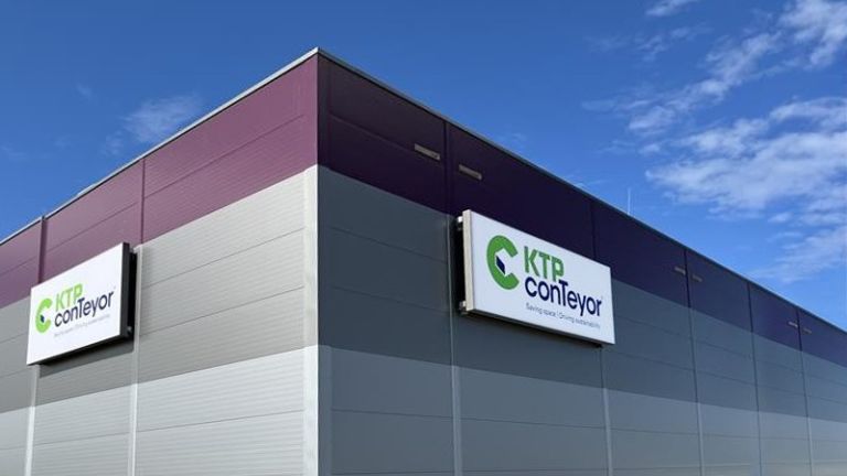 Exterior view of a modern building with a purple and gray facade featuring signs for "KTP Conveyor" on the wall under a blue sky.