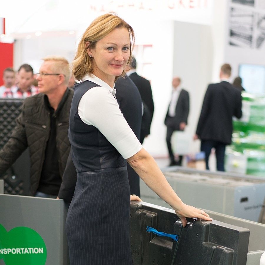 A woman with shoulder-length blonde hair smiles while standing next to a transport box at a busy event, with people in the background.