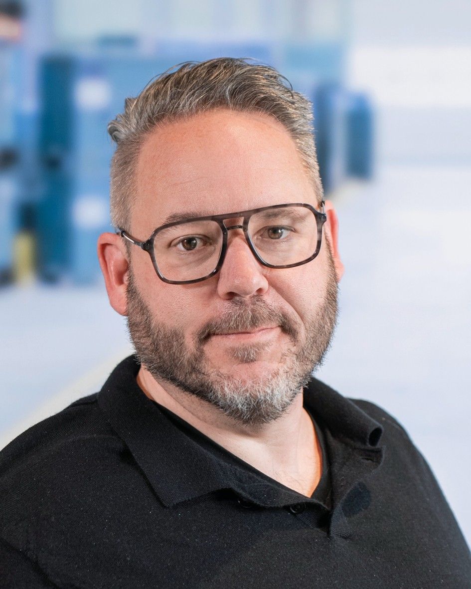 A man with glasses and a beard, wearing a black polo shirt, poses for the camera against a blurred background.