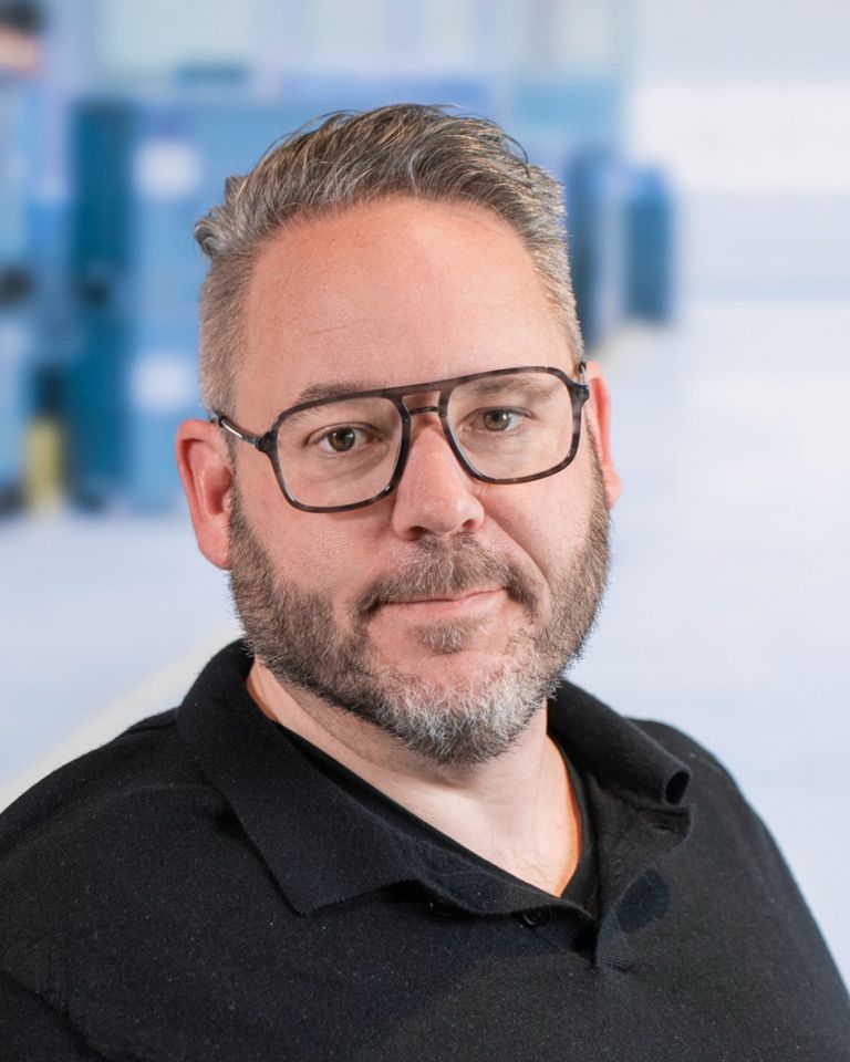 A man with glasses and a beard, wearing a black polo shirt, poses for the camera against a blurred background.