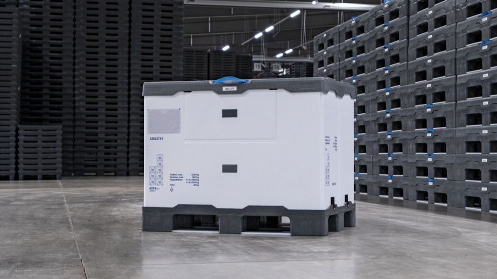 A large white reusable container with a gray lid, positioned on a concrete floor surrounded by stacked black plastic crates in a warehouse setting.