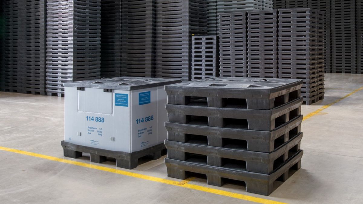 A warehouse interior featuring stacked black plastic pallets and a white container on a concrete floor.