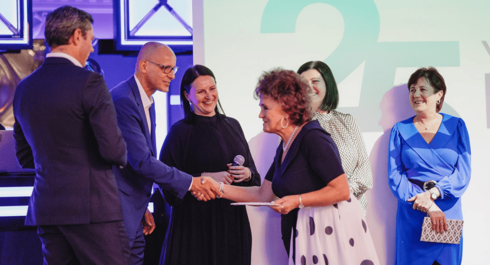 A group of people at a celebration event, with a woman in a polka dot dress shaking hands with a man, while others look on, smiling, against a backdrop with "25 years" displayed.