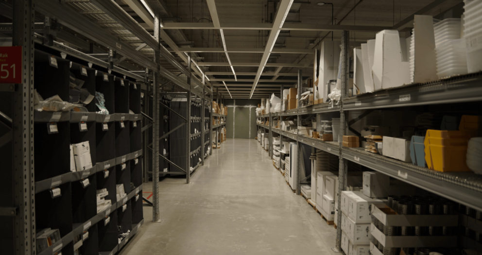 A narrow aisle in a warehouse filled with tall shelves stacked with various products and boxes, under fluorescent lighting.
