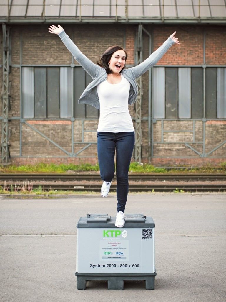 A young woman with brown hair is joyfully jumping on top of a large storage box, with her arms raised in celebration against an industrial backdrop.