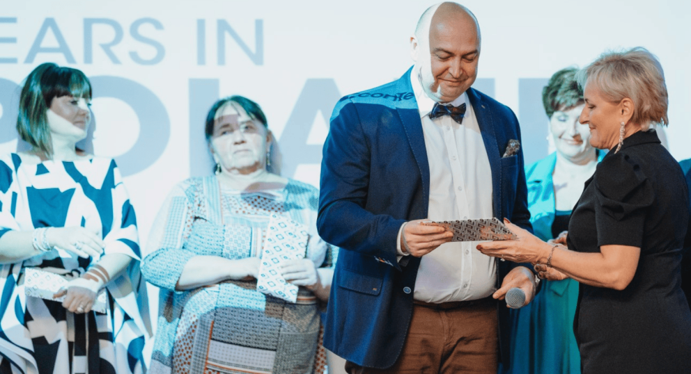 A man in a blue blazer is receiving an award from a woman in a black dress, while several other women stand nearby, and a large "Years in Poland" banner is visible in the background.