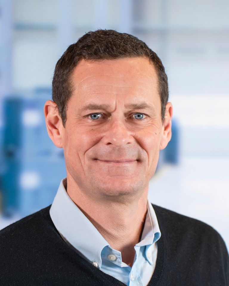 A man with short, dark hair and blue eyes smiles at the camera, wearing a dark sweater over a light blue shirt, with a blurred background.